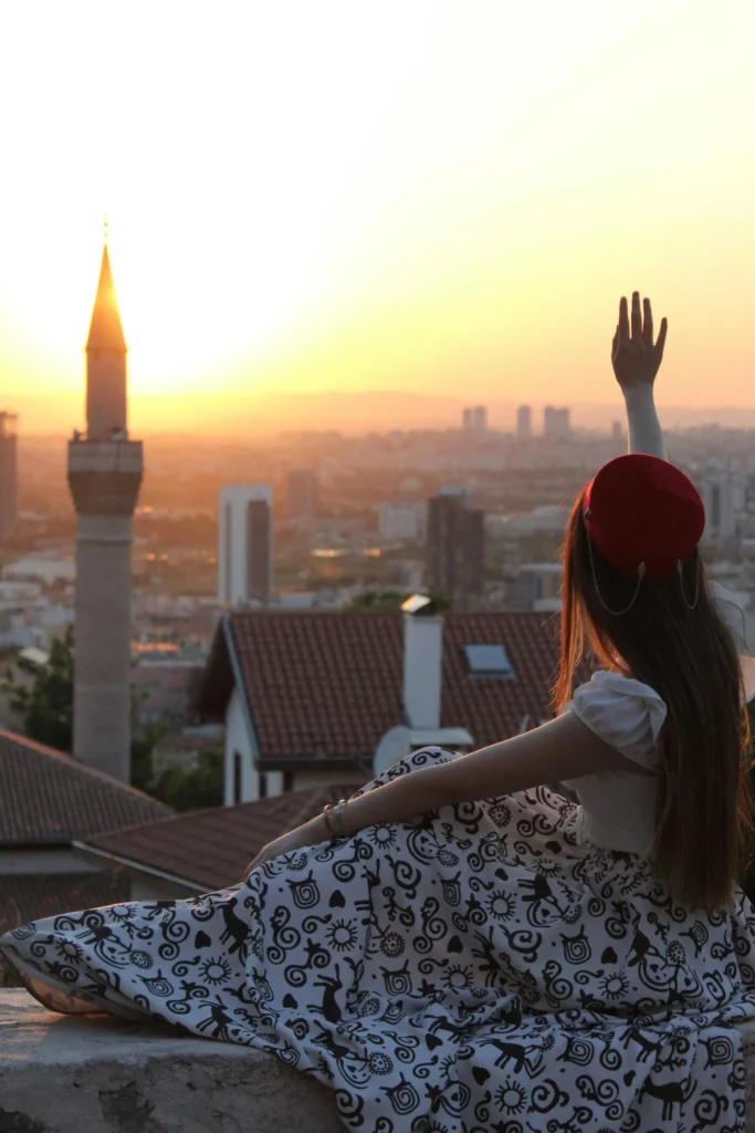  A woman sitting on a rooftop, wearing a patterned skirt and red hat, raising her hand while gazing at a serene sunset over a cityscape.