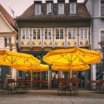 "A bustling city downtown scene featuring a beer garden with parasols, tables, and chairs, set against a vibrant cityscape