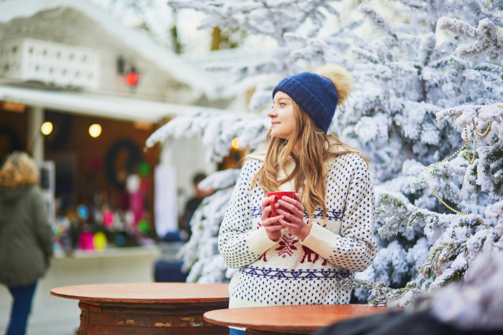 A young woman enjoying a warm drink at a festive Christmas market in Paris in December, surrounded by snow-covered trees and holiday decorations.