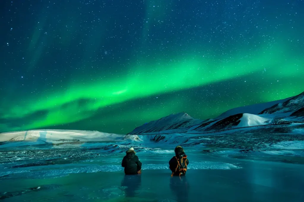 Two people standing under the stunning green aurora borealis in the midnight sky, showcasing the natural beauty of Alaska, USA—one of the best places to live in Alaska.

