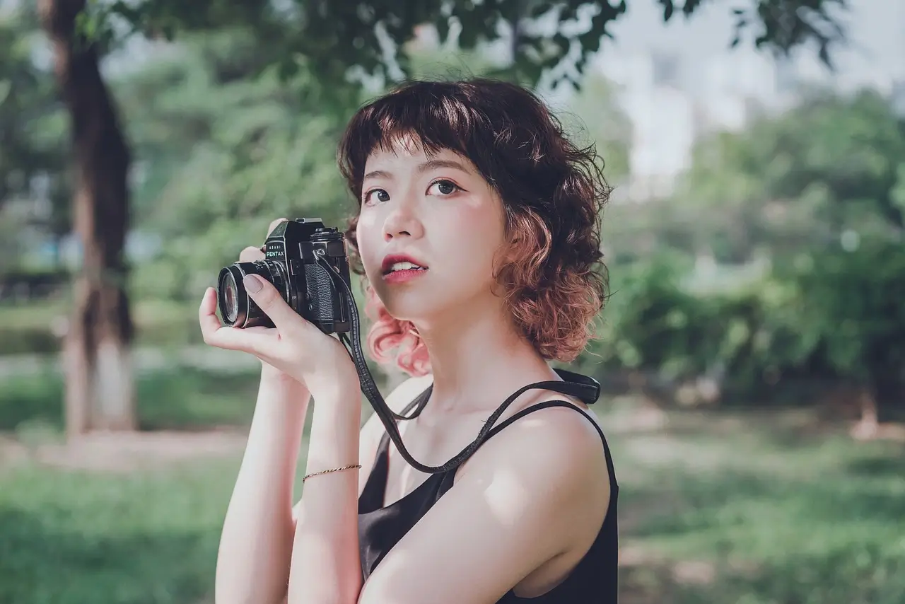 A young woman with a stylish travel aesthetic holding a vintage camera in a lush green park, capturing the beauty of her surroundings.