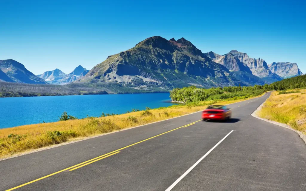 Scenic drive through Glacier National Park in Montana with a red car, blue lake, and dramatic mountain peaks — one of the best places to visit in August USA.