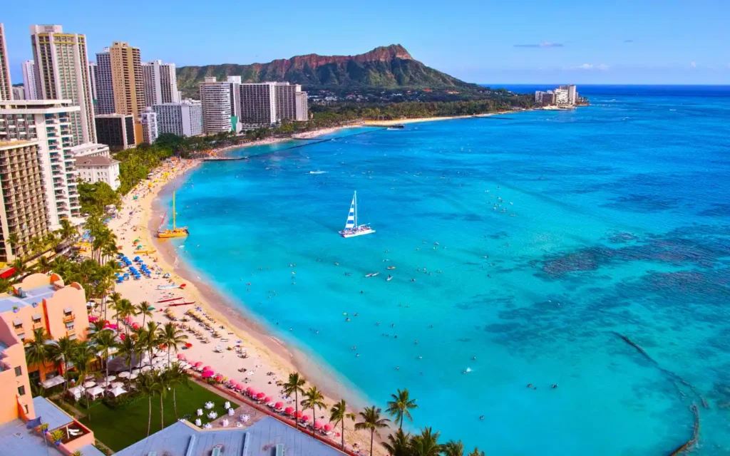 Aerial view of Waikiki Beach, one of the best beaches in Oahu, with palm‑lined golden sand, colorful umbrellas, and a catamaran on turquoise water.